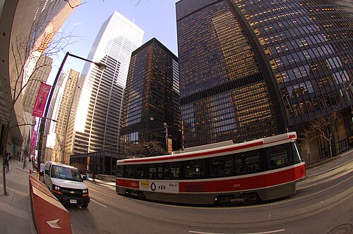A red and white streetcar passing beneath dark glass office towers on a downtown street, a white tower further down the block, the wide lens bending the building lines.