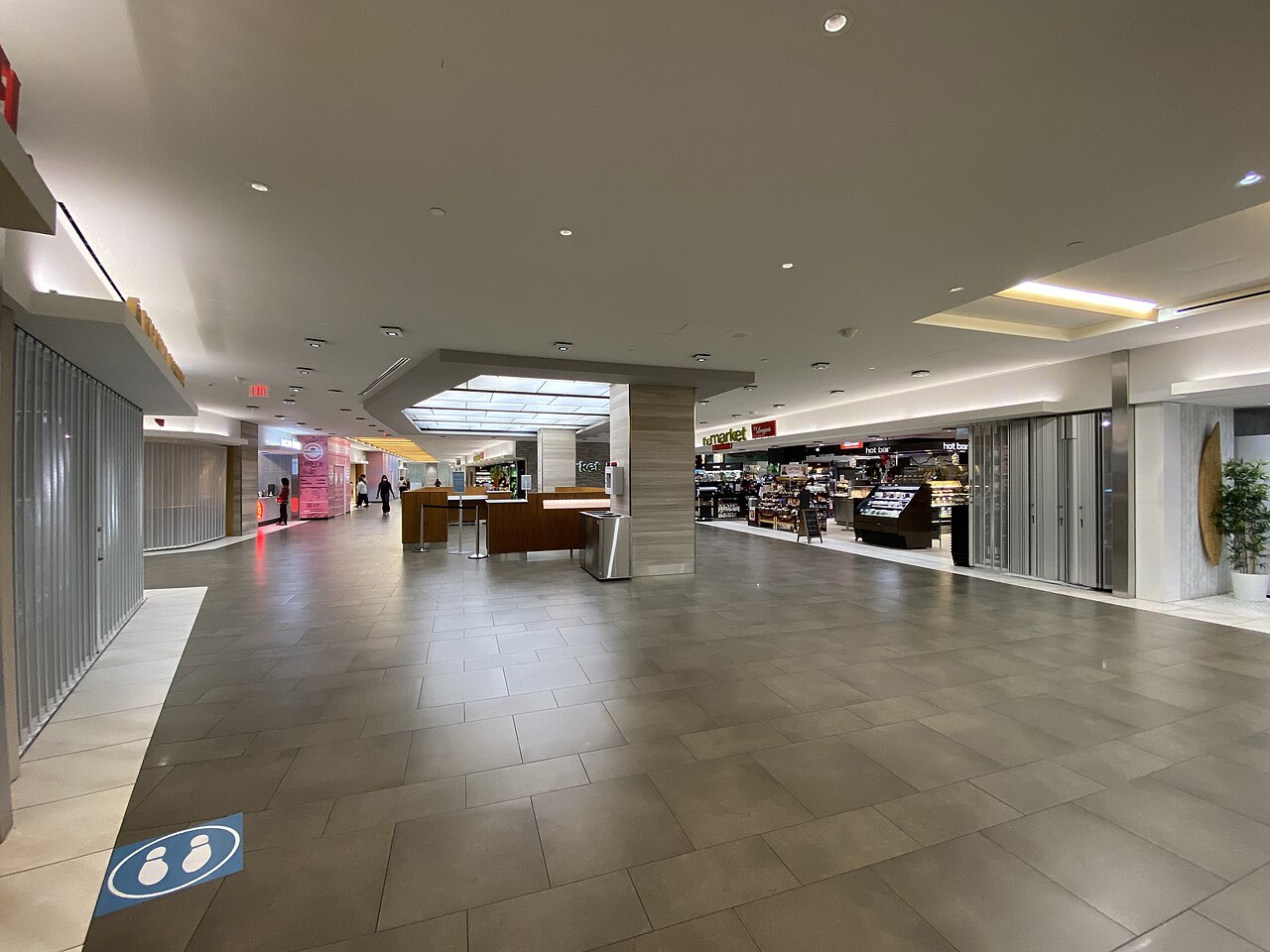 An underground shopping concourse with its storefronts closed behind metal shutters on one side and a small grocery market still open on the other, its hot bar and shelves lit under a low ceiling.