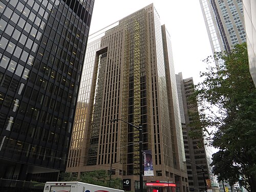 A tan stone office tower with a National Bank branch at street level in red signage, beside a black glass tower, trees along the sidewalk.