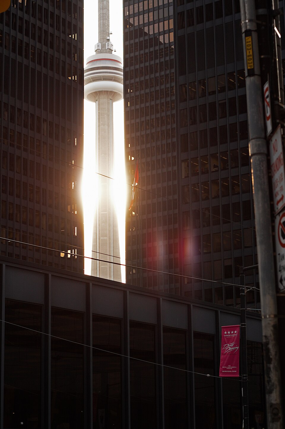 The CN Tower backlit by the sun and framed between two black glass office towers of the Toronto-Dominion Centre, seen from street level.
