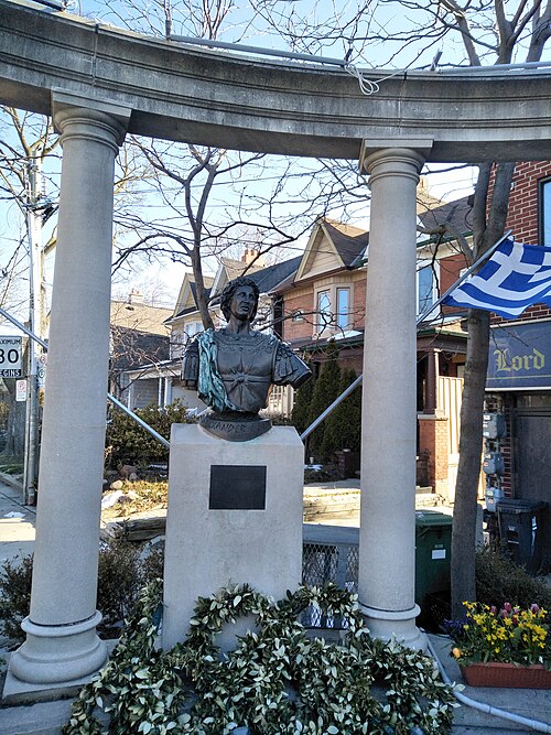 The bronze bust of Alexander the Great at the Alexander the Great Parkette, framed by stone columns, with houses and a storefront sign behind it.
