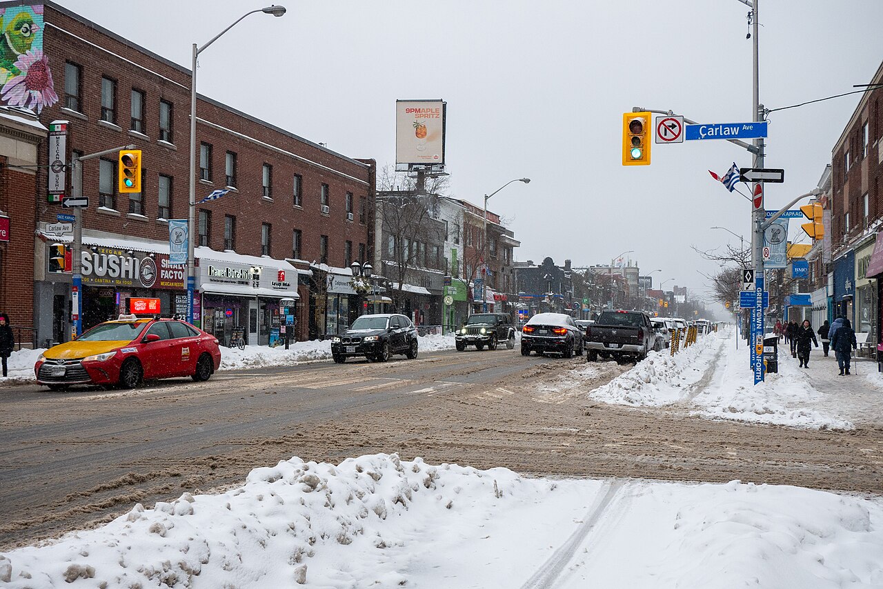 Danforth Avenue looking west toward Carlaw Avenue, storefronts on both sides under snow, cars stopped at a light and pedestrians on the sidewalk.