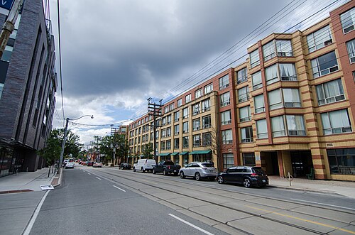 A brick and cream loft style building along Queen Street East, awnings over the ground floor shops and cars parked at the curb under a cloudy sky.