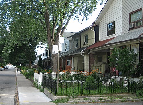 A row of two storey wood frame houses on a residential street, small iron and wood fences around the front gardens and a large tree over the sidewalk.