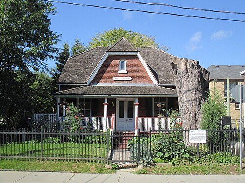 Maple Cottage, a red brick house with a steep gambrel roof and a small sign over the porch, the wide stump of the Maple Leaf Forever tree standing beside it behind an iron fence.