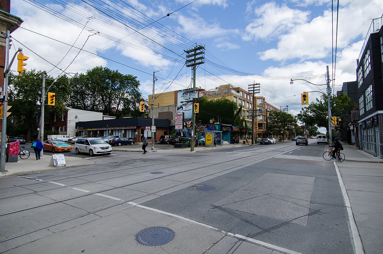 Queen Street East at a wide intersection, low storefronts on both sides, streetcar tracks down the middle and a cyclist crossing at a green light.