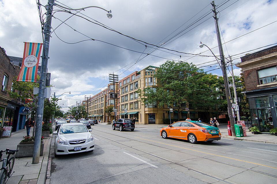 Queen Street East between Winnifred and Caroline Avenues, storefronts and a loft building along the block, a banner reading Leslieville on a lamppost and a taxi waiting at the light.