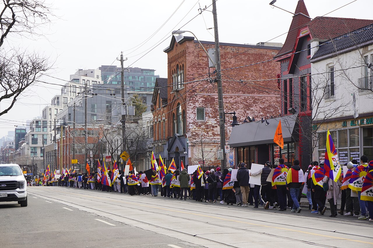 A march carrying Tibetan flags down Queen Street West, storefronts and a red brick Victorian building with a turret along the sidewalk.