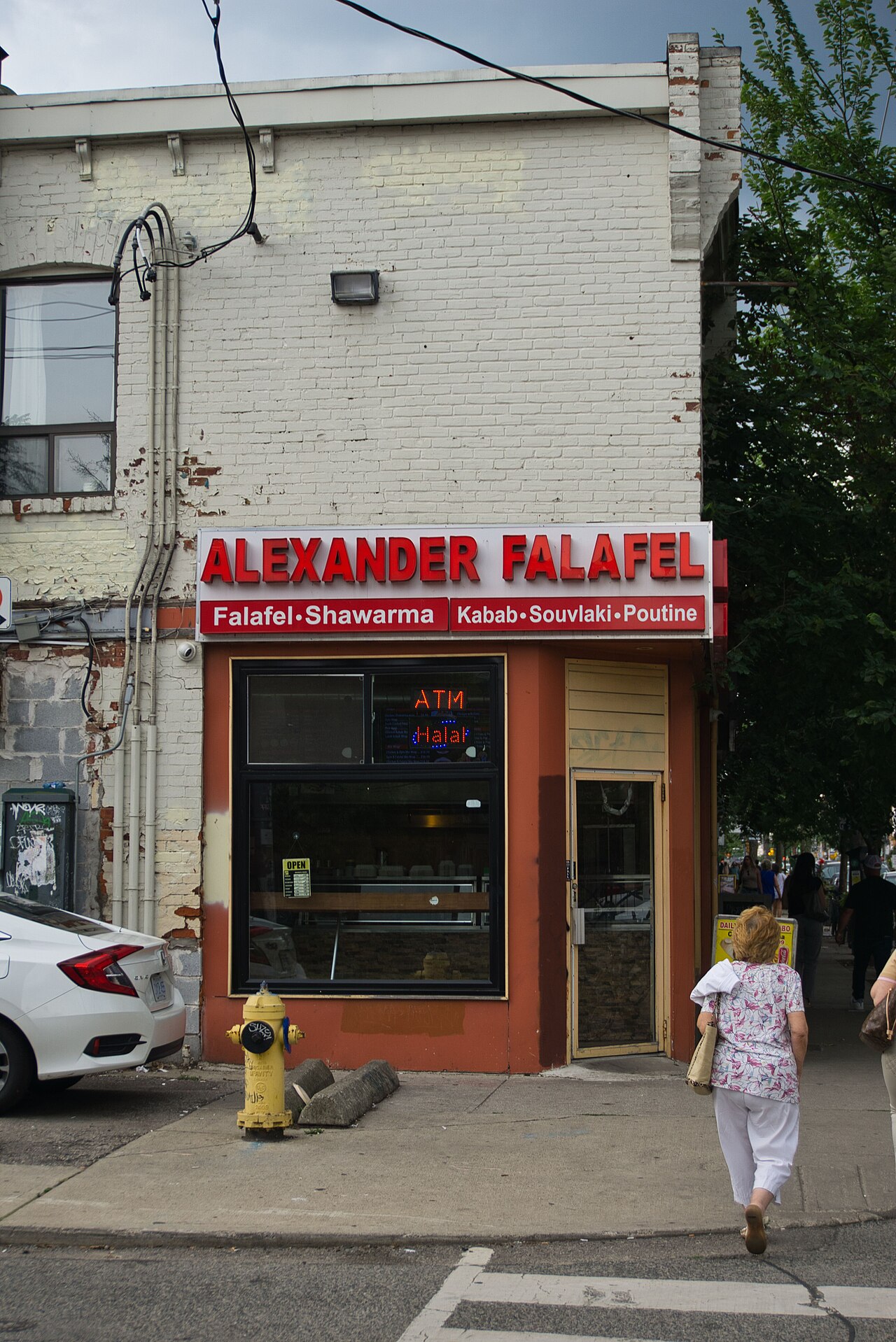 Alexander Falafel's corner storefront in whitewashed brick, red signage over the windows and a yellow fire hydrant on the sidewalk.
