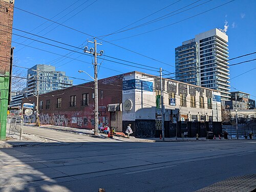 A vacant storefront building at Noble Street on Queen Street West, boarded windows covered in graffiti and For Lease signs, condo towers rising behind it.