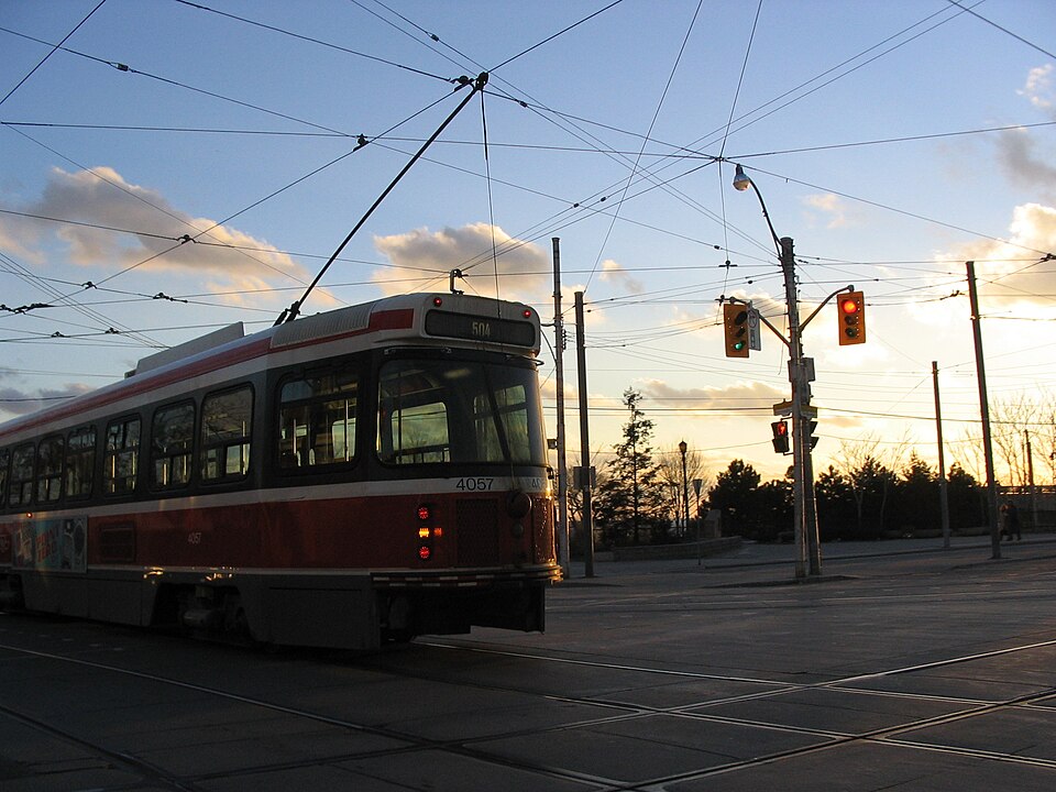 A TTC streetcar stopped at Queen Street West and Roncesvalles Avenue at dusk, overhead wires crossing above the intersection and traffic lights turning red.