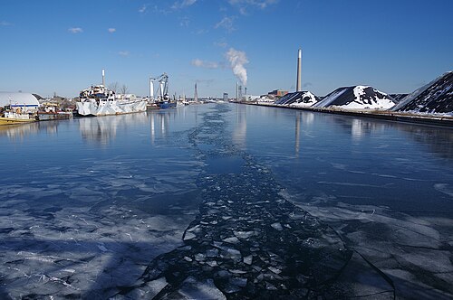 The ship channel frozen over in winter, a support vessel and crane barge moored on the left, mounds of salt and aggregate piled along the right bank and a smokestack visible in the distance.