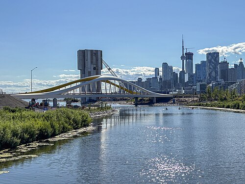 A newer white and yellow arched bridge carrying Cherry Street over the water, construction equipment on the near bank and the downtown Toronto skyline and CN Tower behind it.