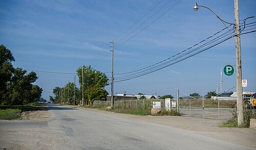 Unwin Avenue lined with chain link fencing and parked equipment, a parking sign mounted on a utility pole and the CN Tower visible far down the road.