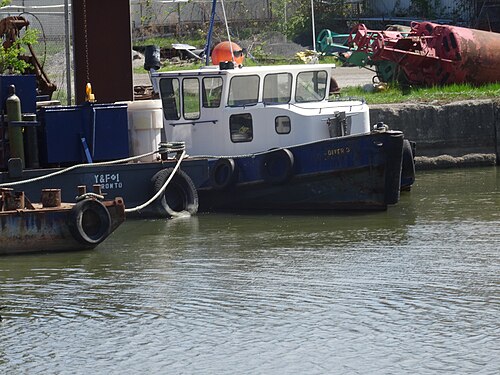 The tugboat Diver 3 and a companion barge moored at a dock along the channel, old tires used as fenders and salvage equipment stacked on the bank behind them.