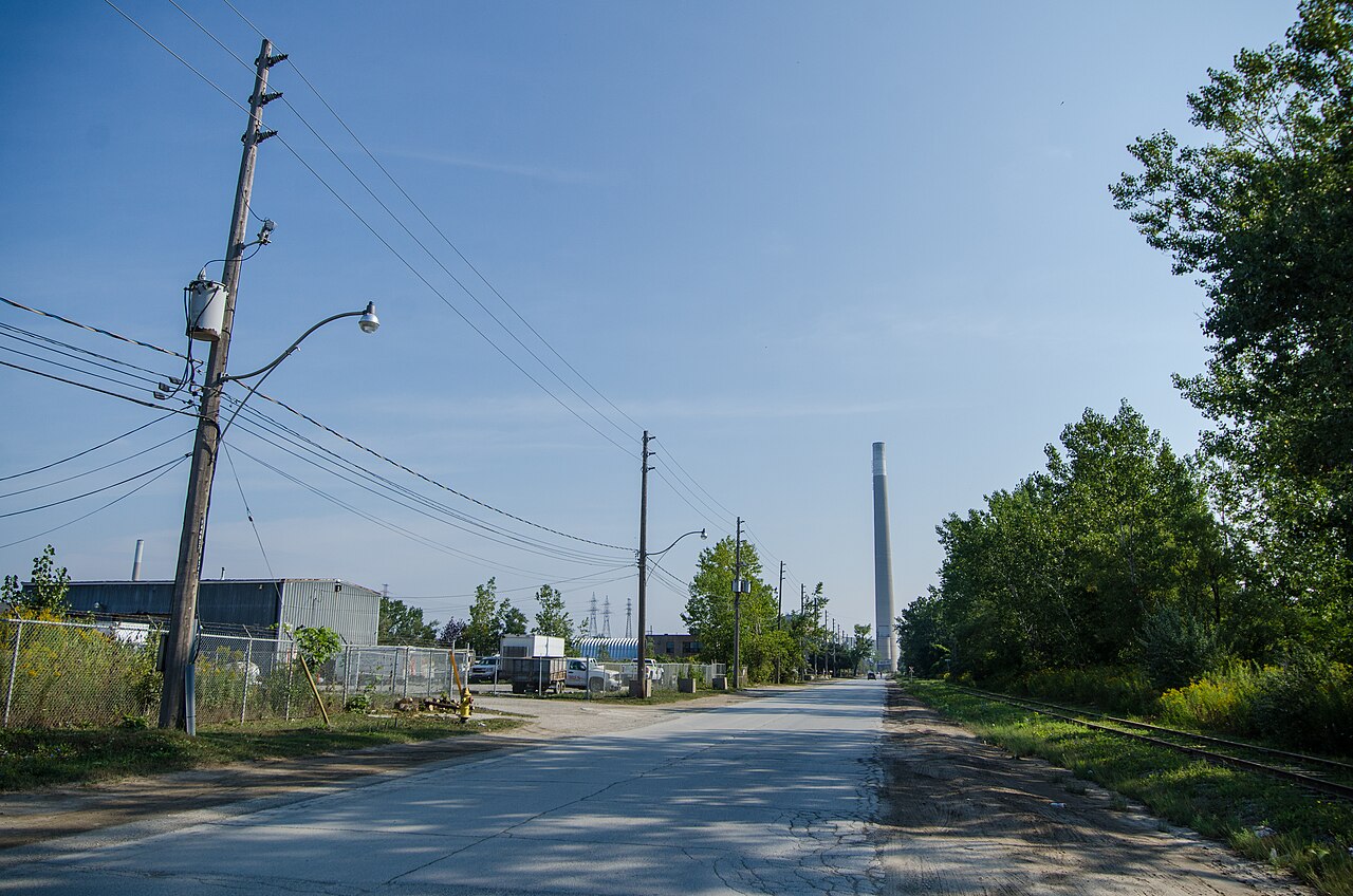 Unwin Avenue, a wide gravel shouldered road running straight toward the Hearn Generating Station's smokestack, a rail spur along the right side and a small industrial shed behind a chain link fence.