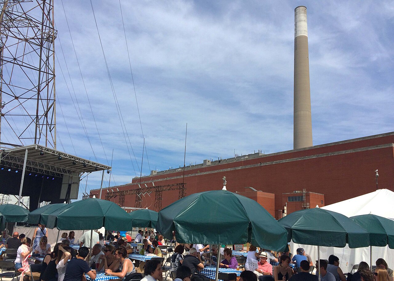 The Hearn Generating Station's red brick wall and single tall smokestack, seen from a public event held on the site, string lights and tent umbrellas in the foreground.