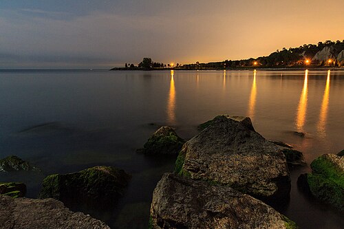 Lake Ontario at dusk, mossy boulders in the foreground and the pale bluffs lit by shoreline lights reflecting across calm water in the distance.