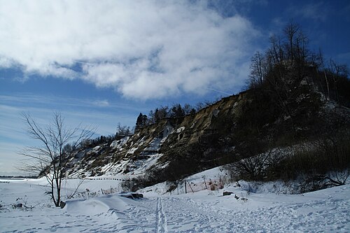 The bluffs in winter, snow covering the frozen shoreline below a tree covered cliff face streaked with exposed clay.
