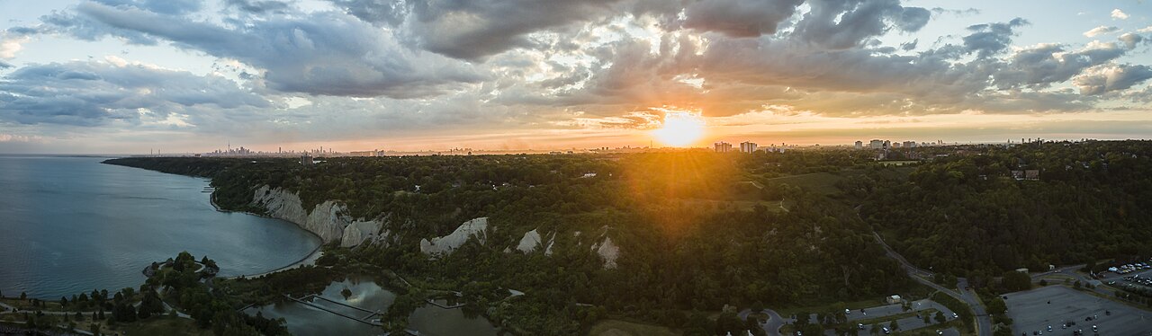An aerial view above the Scarborough Bluffs at sunset, the pale cliff line curving away along Lake Ontario with the Toronto skyline visible in the distance and Bluffer's Park's marina and parking lot at the foot of the slope.