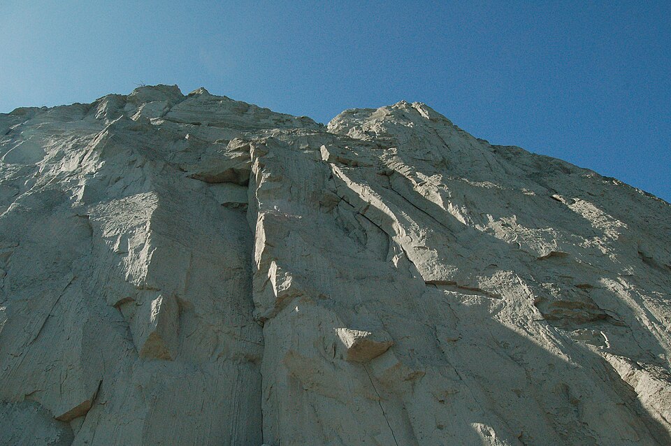 Looking straight up a sheer, sunlit wall of the bluffs, the pale clay cut through with vertical fissures and a slide of loose material at its base.