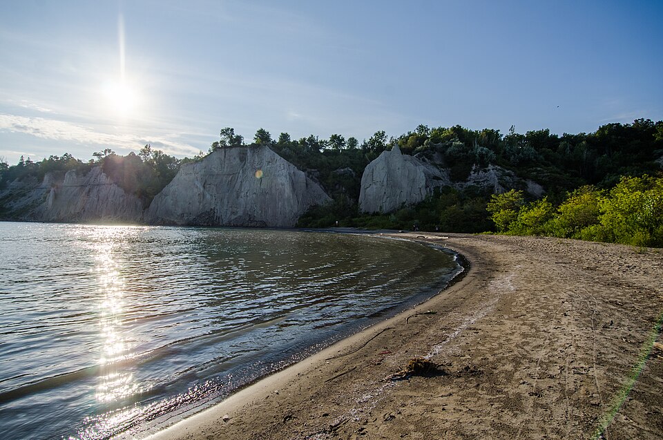 Pale eroded cliffs rising straight from a curved sand beach at Bluffer's Park, two pillars of clay standing apart from the main cliff face, the water pale green near the shore.