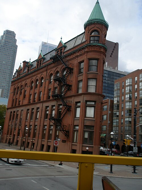 The Gooderham Building's rounded corner, a copper topped turret and a zigzag fire escape rising above Wellington Street, taller Financial District towers behind it.