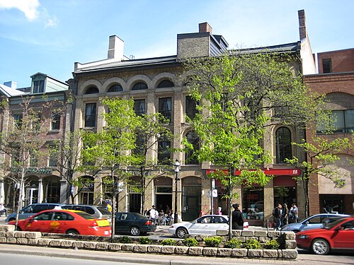 A row of nineteenth century buildings on Front Street, ground floor cafes under arched third story windows, trees and parked cars along the curb.