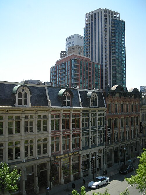 A mansard roofed row of Front Street buildings seen from above, Europe Bound and Winners storefronts at street level and condo towers rising behind them.