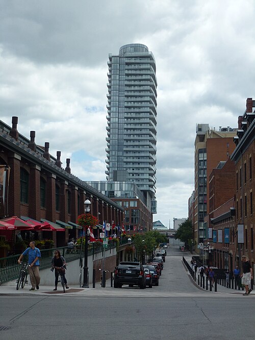 Market Street looking south from Front Street, St. Lawrence Market's dormered roofline on one side and a glass condo tower rising at the far end, cyclists on the sidewalk.