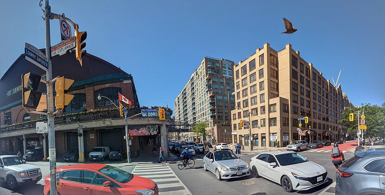 The corner of Jarvis and Front Streets, St. Lawrence Market's brick facade and sign on the left, traffic waiting at the lights and a bird flying over the intersection.