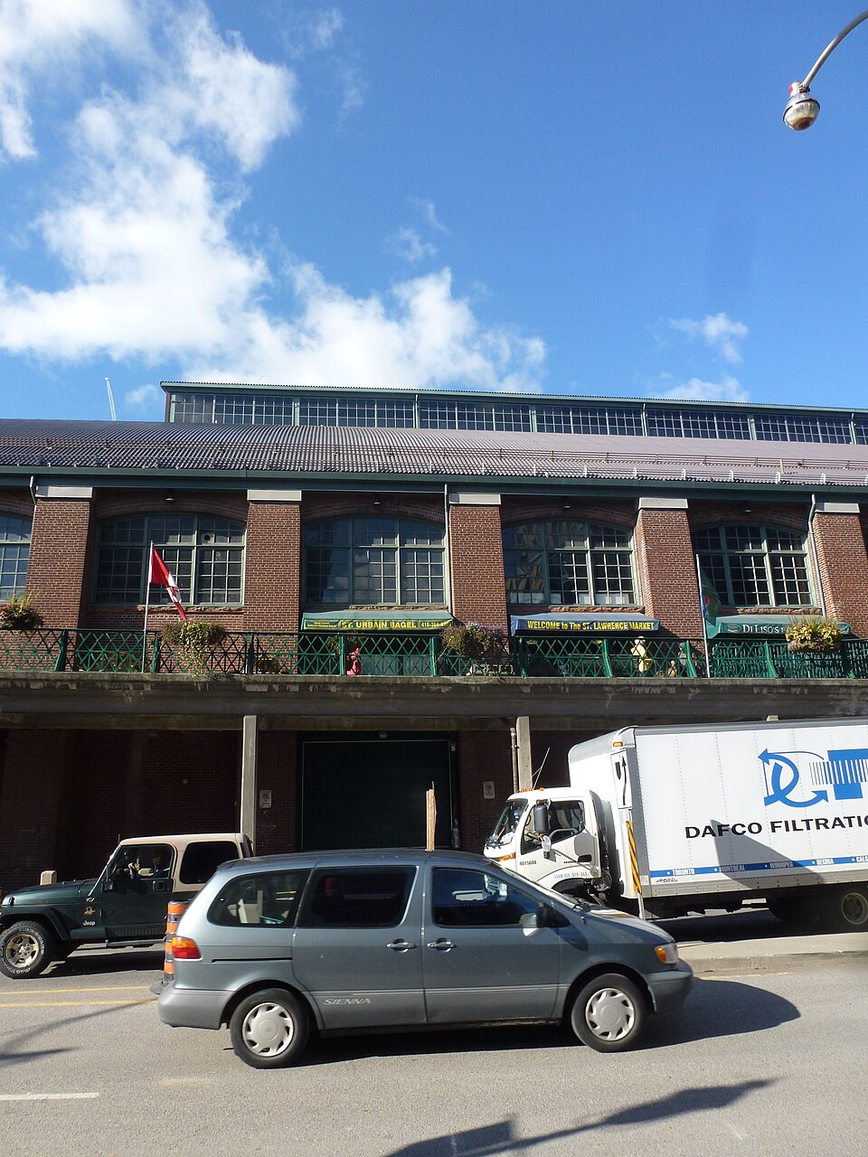 St. Lawrence Market's eastern facade, arched windows above a green iron balcony strung with a Welcome to St. Lawrence Market banner, a delivery truck parked below.