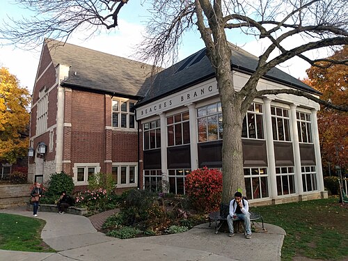 The Beaches Branch of the Toronto Public Library, a brick building with a colonnaded reading room window, seen across its garden with someone sitting on a bench under the tree out front.