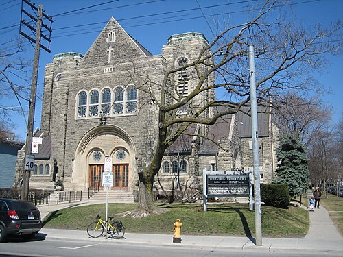 Corpus Christi Catholic Church, a grey stone Gothic Revival building with a rose window and a wide arched entrance, seen from across Queen Street East.