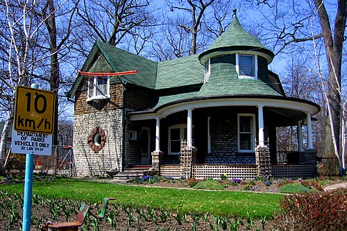 A Queen Anne style house near Kew Gardens with a wraparound porch and a round turret under a bell shaped green roof.