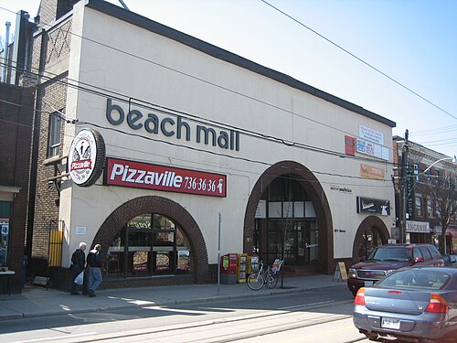 A cream stucco building with three arched brick entrances on Queen Street East, now a small mall, with a pizza shop under one of the arches.
