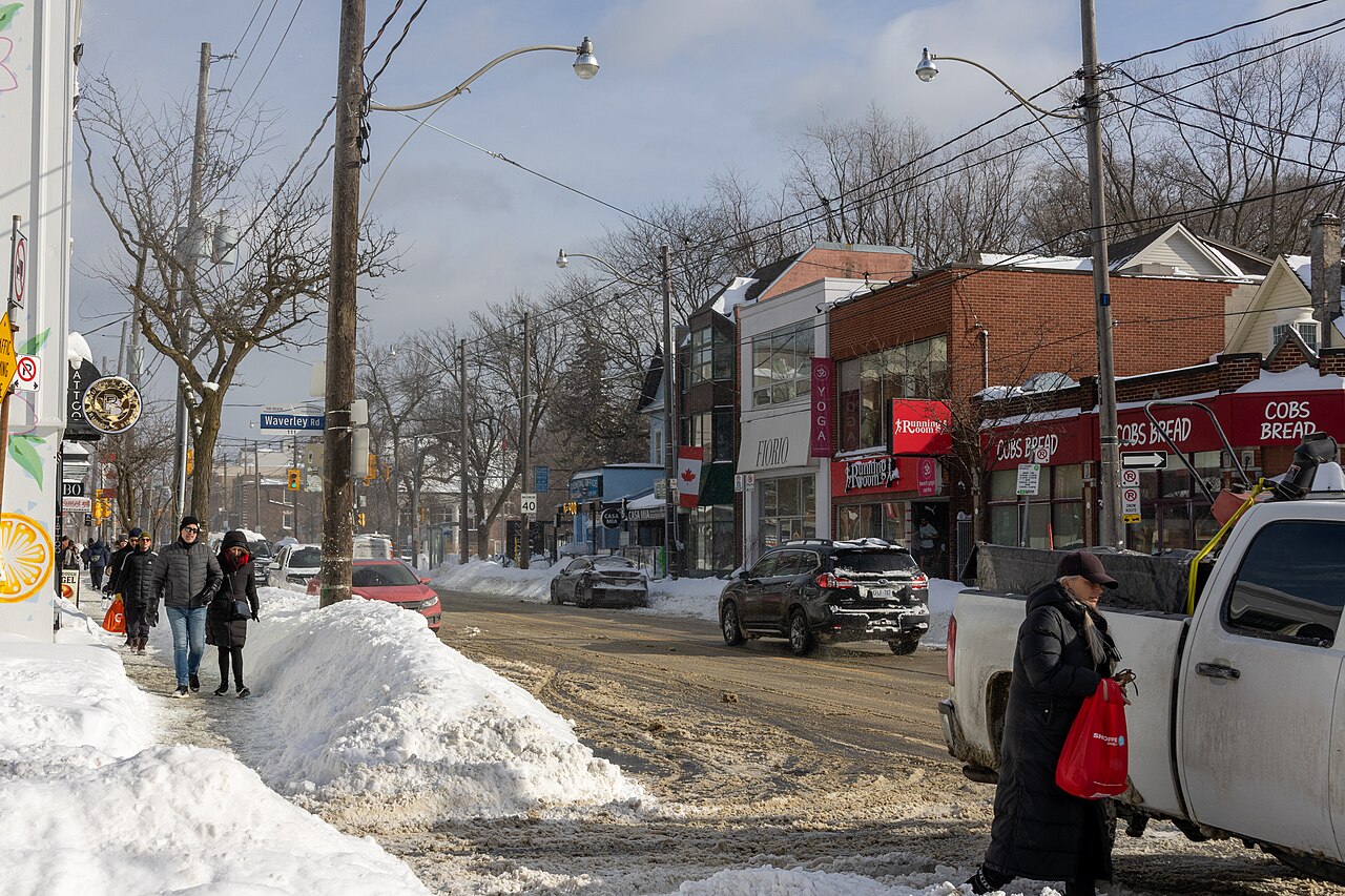 Queen Street East in winter near Waverley Road, snow banked along the curb, storefronts including a bakery and a yoga studio, and a pickup truck stopped in the road as pedestrians pass on the sidewalk.