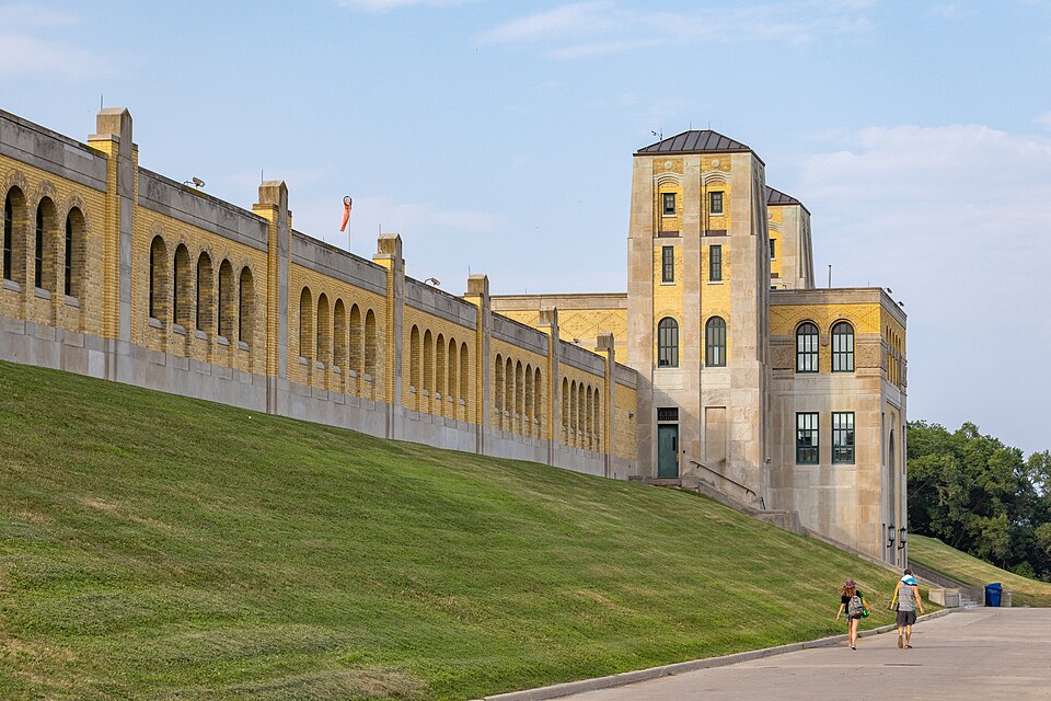 The plant's long arcaded wall and central tower in yellow brick and stone, seen from the sloped lawn below with two people walking up the path.