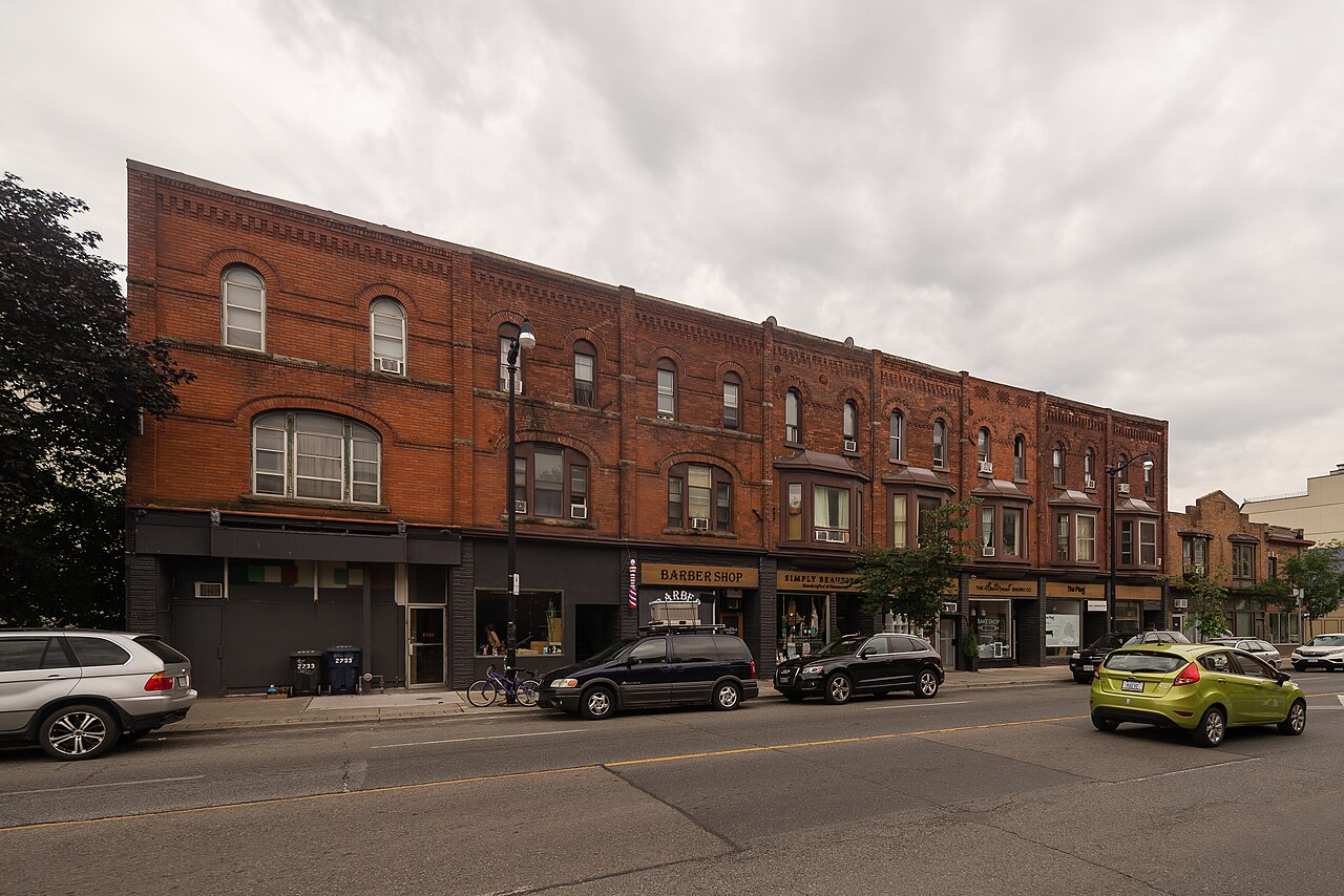 Dundas Street West, a run of two and three storey Victorian brick storefronts with round arched upper windows, cars parked along the curb in front of a barber shop and a beauty salon.
