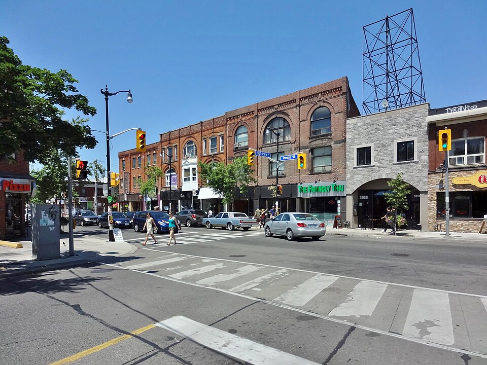 The corner of Dundas Street West and High Park Avenue, a lattice steel water tower rising behind the roofline, pedestrians in the crosswalk and a pizza shop sign at the corner.