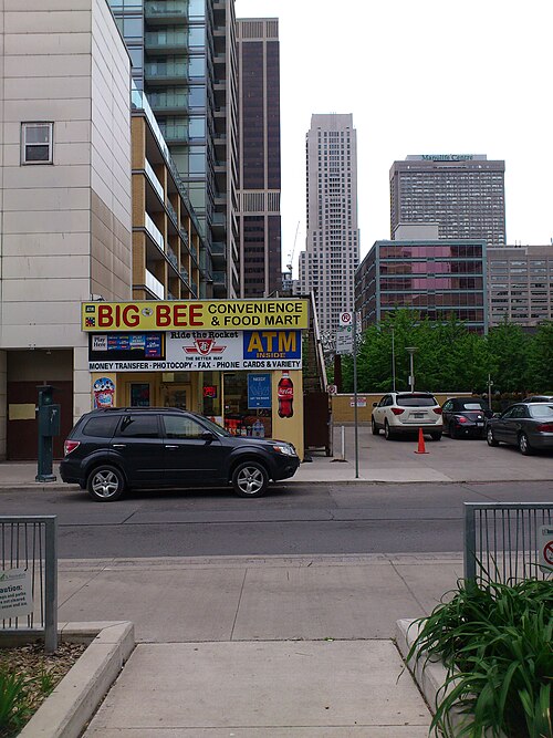 A small convenience store on a corner beside a parking lot, condo and office towers rising behind it.