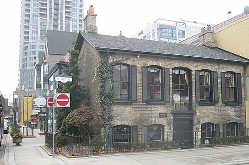 A small stone and brick house on Yorkville Avenue, dwarfed by the glass and brick towers behind it.