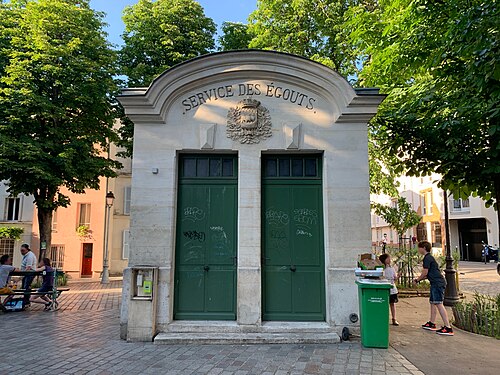 The stone building of the Paris sewer service on place du Guignier, two tall green doors under SERVICE DES ÉGOUTS carved above them with the city's arms. People are sitting at a table under the trees on the left.