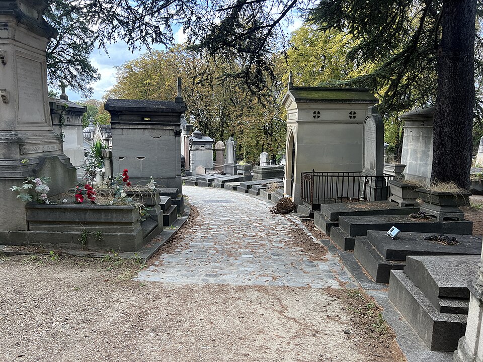 Chemin du Tertre inside the cemetery, a narrow cobbled path bending between stone family chapels, with a cedar branch over it and an iron railing round one of the plots.