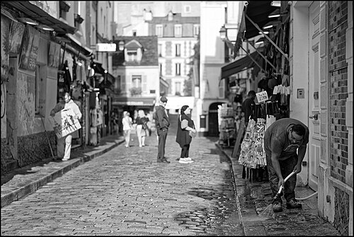 A cobbled street of souvenir shops opening up in the morning, with one man hosing out the gutter and another carrying a framed canvas past the awnings.