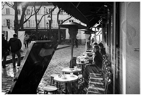 A café terrace on Place du Tertre, gas heaters between the marble tables and rattan chairs along the window, with the Galerie Montmartre shopfront across the square.