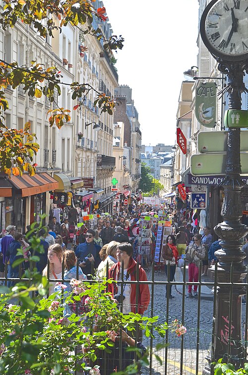 Rue de Steinkerque seen down the hill from Place Saint-Pierre, both pavements taken up by souvenir racks and clothes rails, a cast iron street clock and a tabac sign on the right.