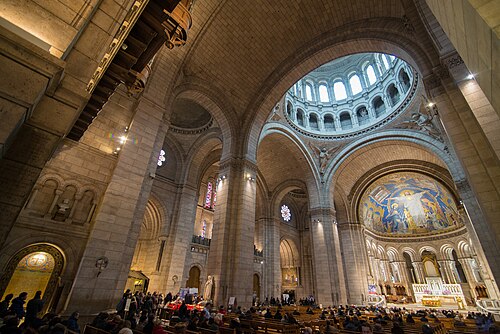 Inside the basilica, looking across the crossing towards the apse, with daylight coming down through the dome and the pews full.