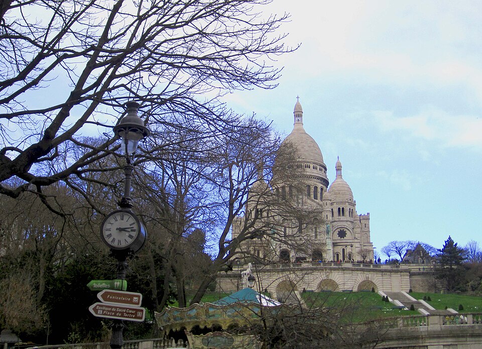 The bottom of Square Louise-Michel in winter. The roof of the carousel is in the foreground, a lamp post has direction signs for the funicular and Place du Tertre, and the stone terraces and steps run up to the basilica.