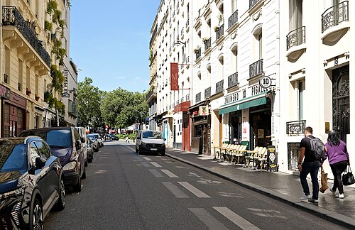 Rue Frochot looking toward Place Pigalle, a hotel sign on the left, a bar with a green awning and pavement chairs on the right, and the trees of the square at the far end.